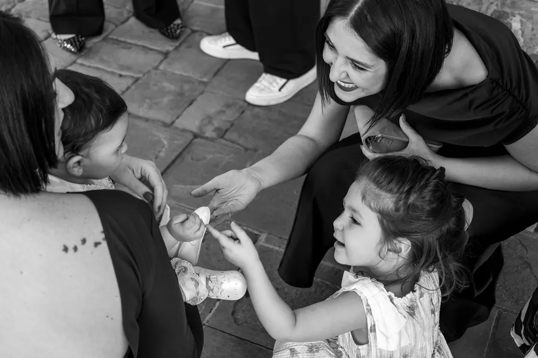 In bianco e nero, una bimba porge qualcosa al bebè tenuto in braccio, mentre una donna si china sorridendo.