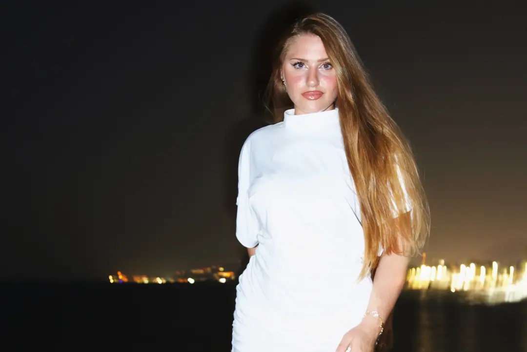Una ragazza in bianco sulla riva, con le luci della costa sfocate alle spalle.