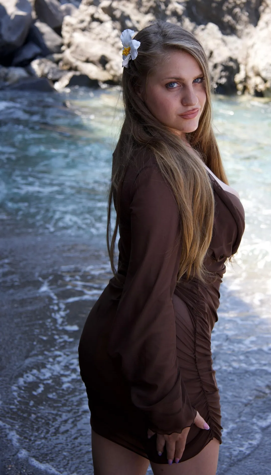 Di spalle, con un abito marrone e un fiore bianco tra i capelli, guarda in camera vicino al mare.