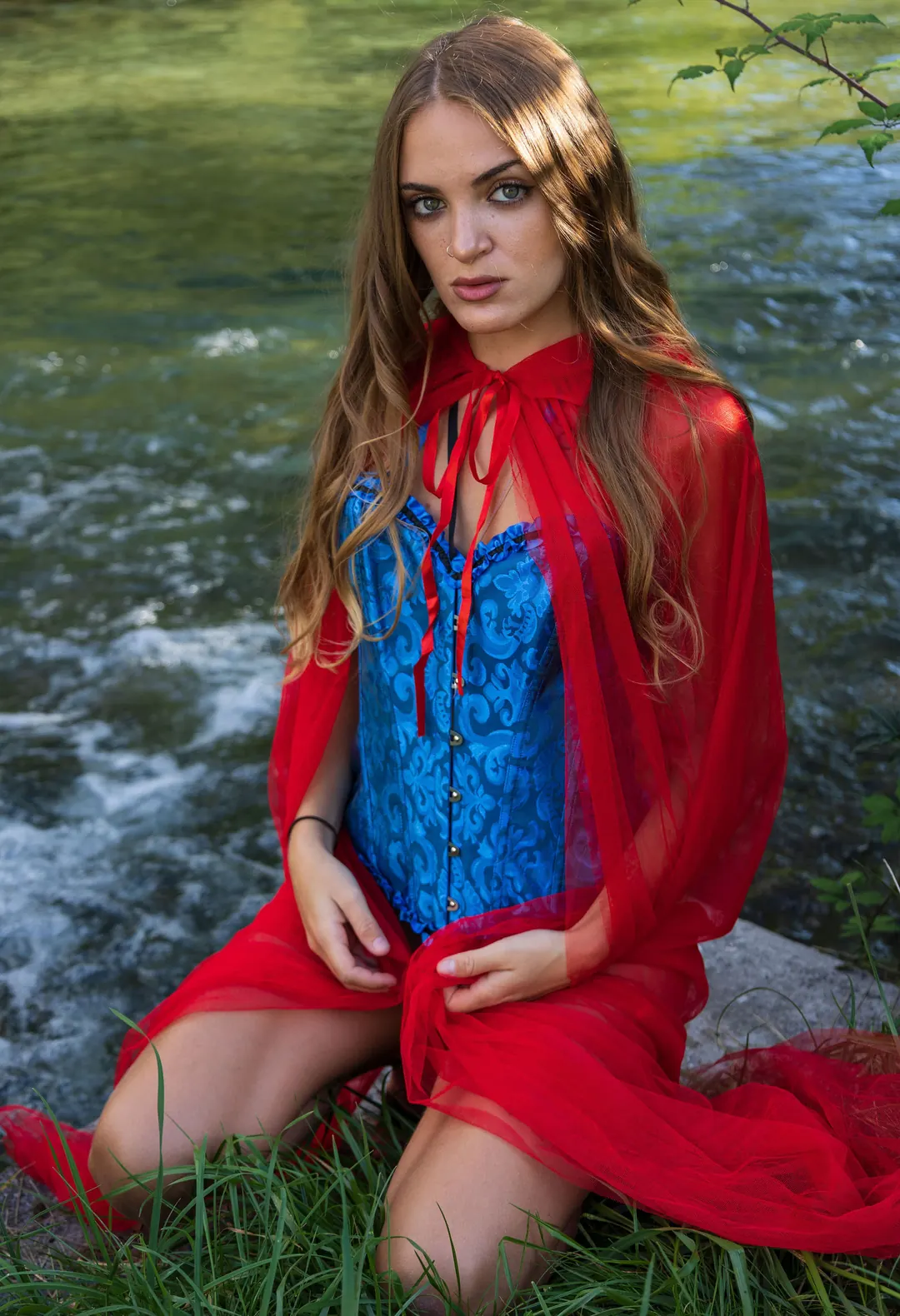 Una ragazza con il mantello rosso e il corsetto blu, inginocchiata sull'erba vicino al torrente.